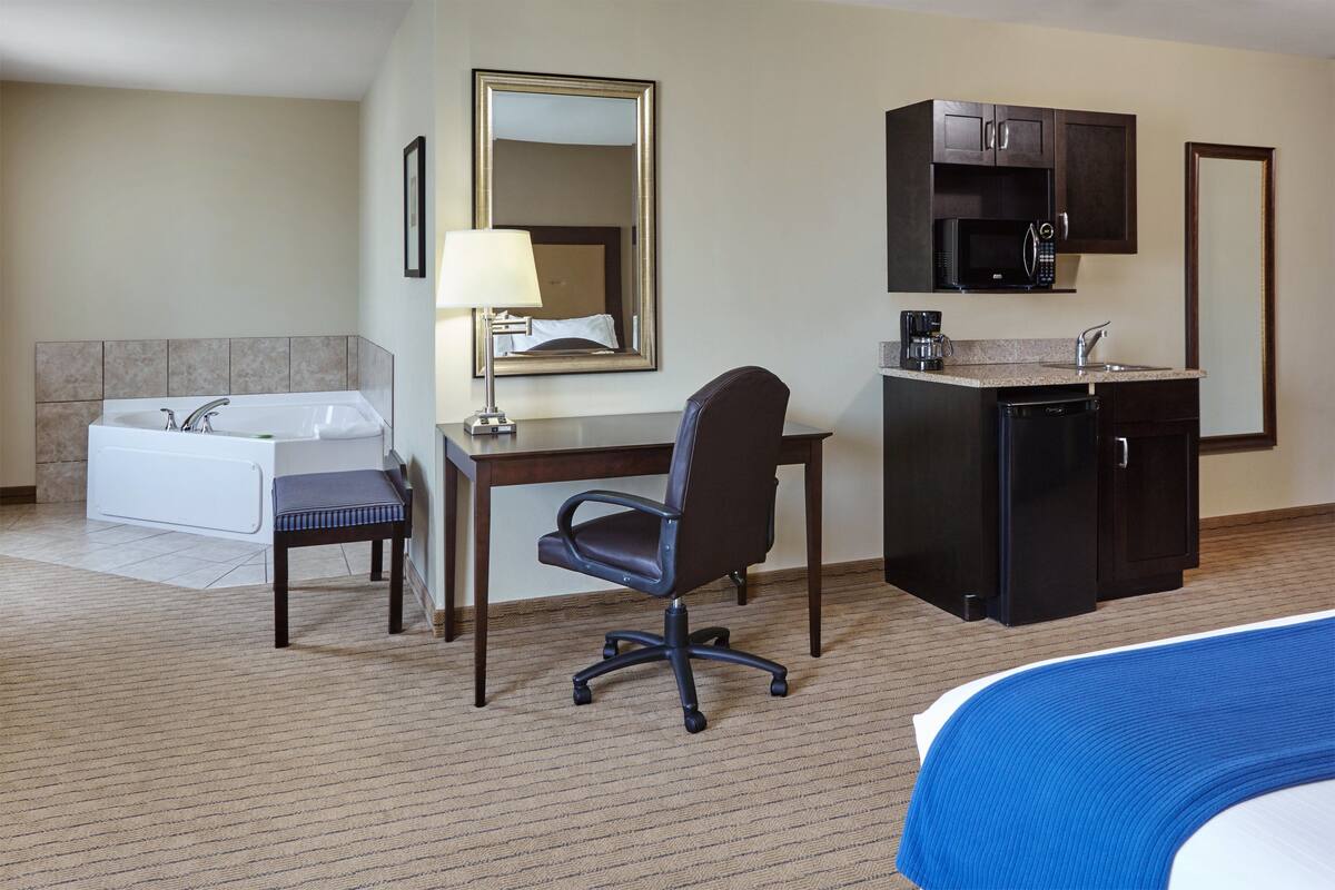 A hotel room with a bed, a desk with a chair, a small kitchenette, a bathroom with a bathtub, and a large mirror.