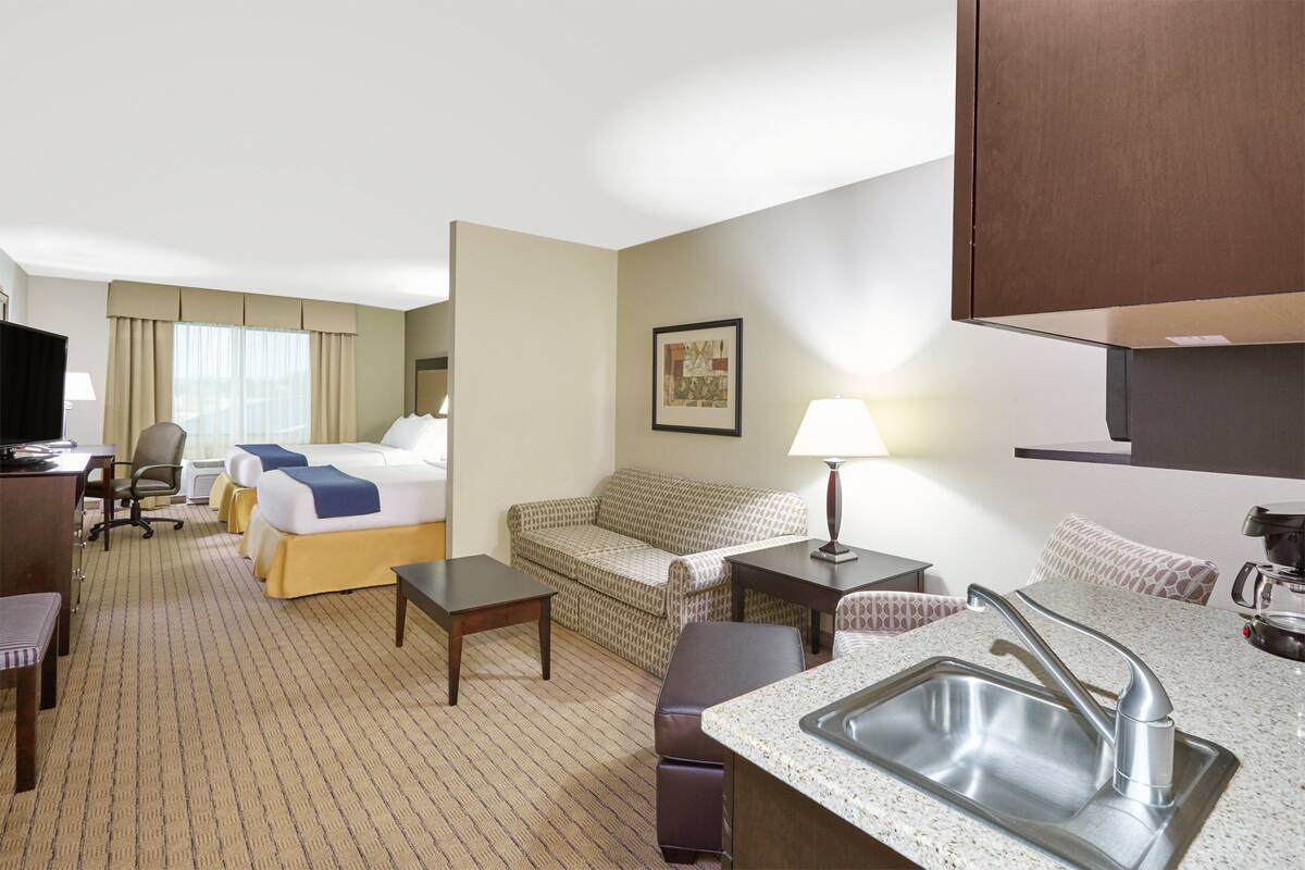 A hotel room with a bed, a sofa, a desk, a chair, a small table, a sink, and a television.