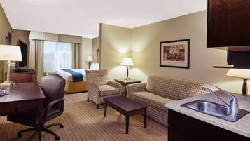 Une chambre d’hôtel comprenant un lit, un canapé, un bureau avec une chaise, une petite table, une lampe, un tableau au mur et un lavabo.