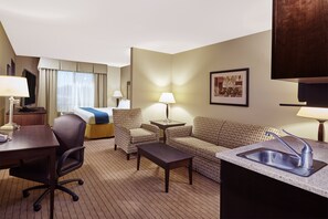 Une chambre d’hôtel comprenant un lit, deux fauteuils, une table basse, un bureau, une lampe, un tableau au mur et un lavabo.