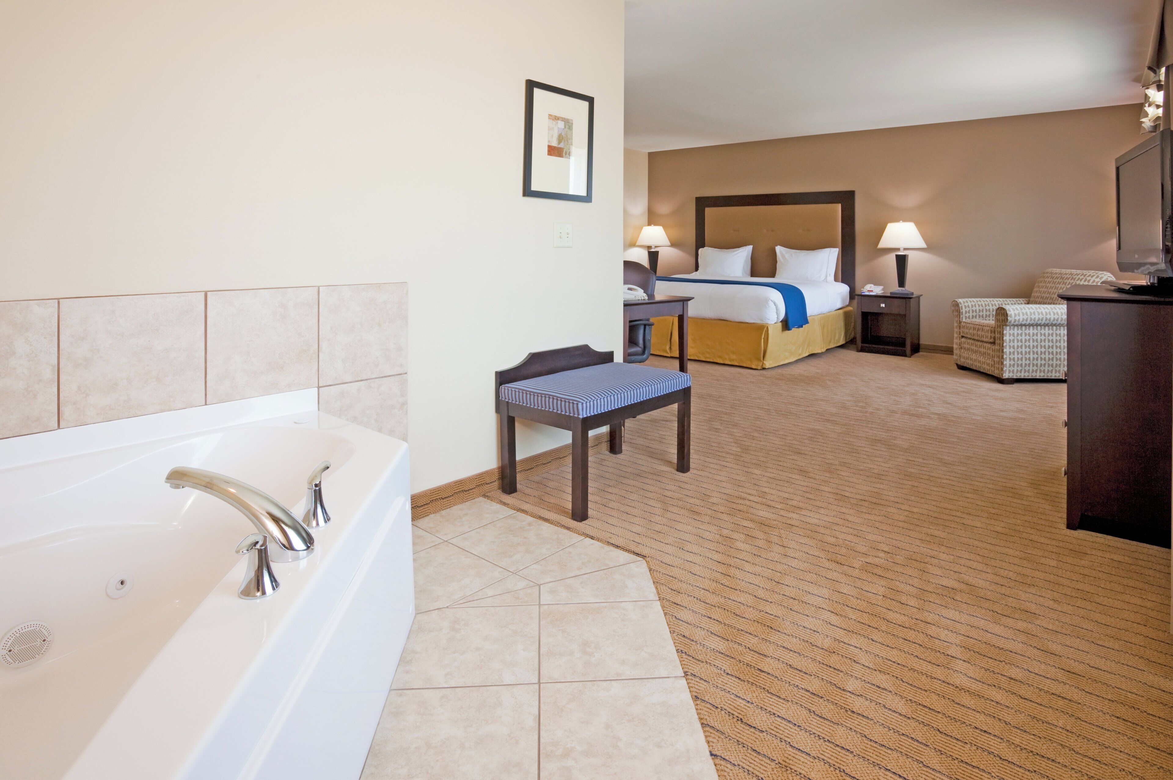 A hotel room with a large bathtub, a bed with a headboard, a bedside table with a lamp, and a small table with a chair.