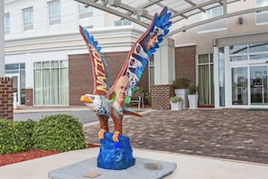 A sculpture of a bird with wings, placed on a concrete base, in front of a building with a brick facade and large windows. - Holiday Inn Statesboro-University Area by IHG (Statesboro)