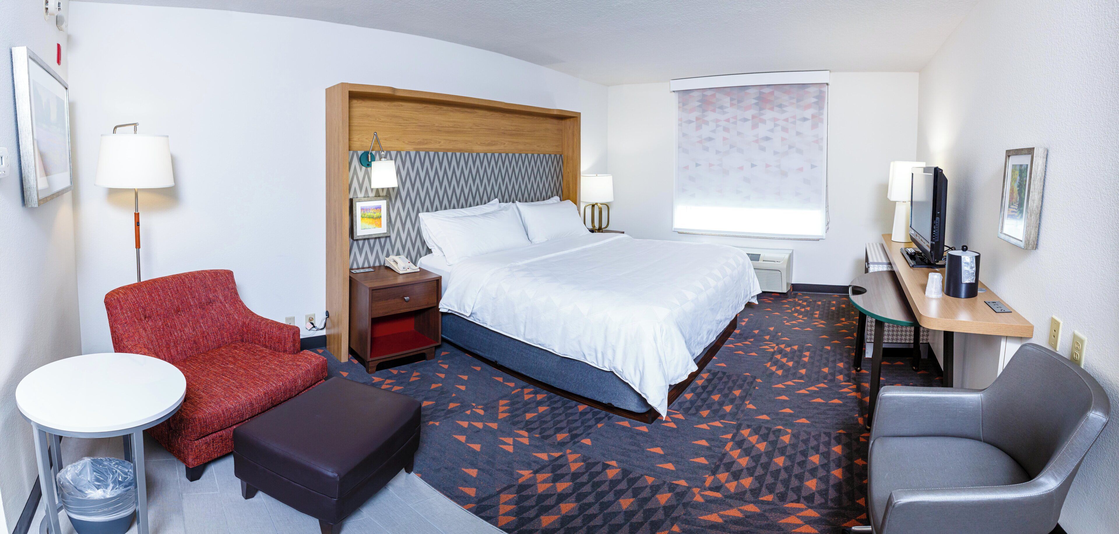 A hotel room with a bed, a desk, a chair, a small table, and a trash bin.