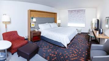 Une chambre d’hôtel avec un lit, un bureau avec un ordinateur, une chaise, une petite table et un fauteuil rouge.