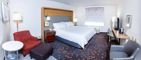 A hotel room with a bed, a desk with a computer, a chair, a small table, and a red armchair.