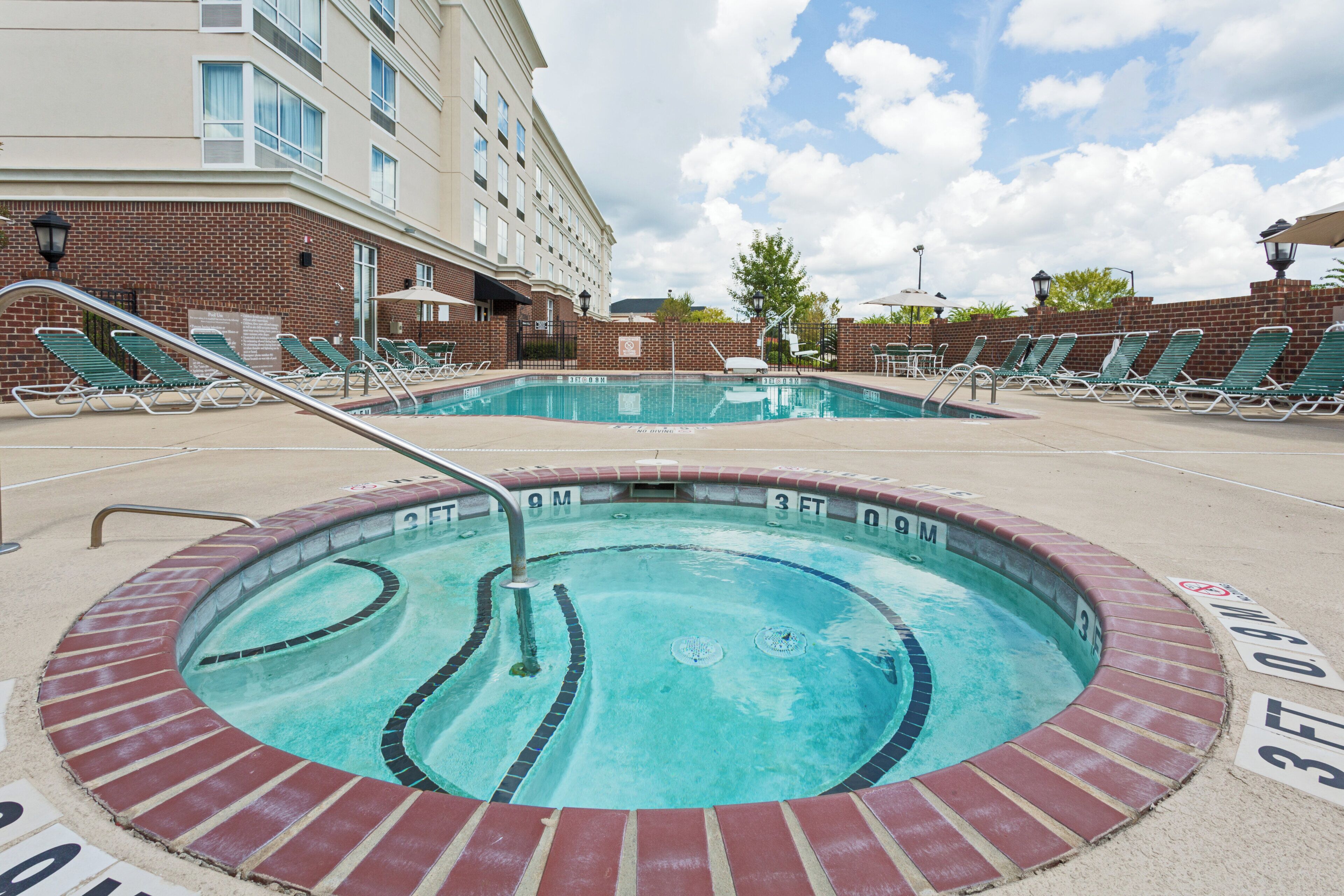 Une piscine avec un jacuzzi, entourée de chaises longues et bordée d’un bâtiment en briques.