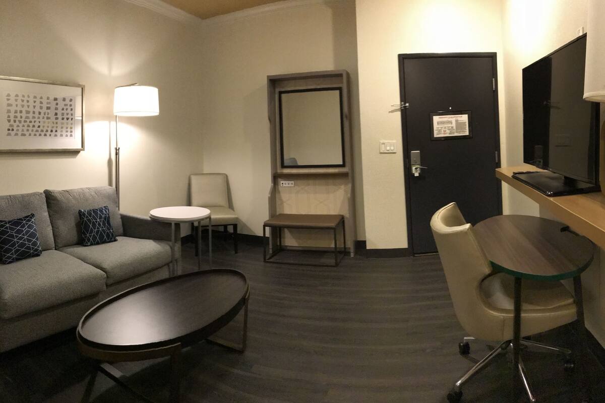 A hotel room with a grey sofa, a small round table, a desk with a chair, a TV, and a lamp.