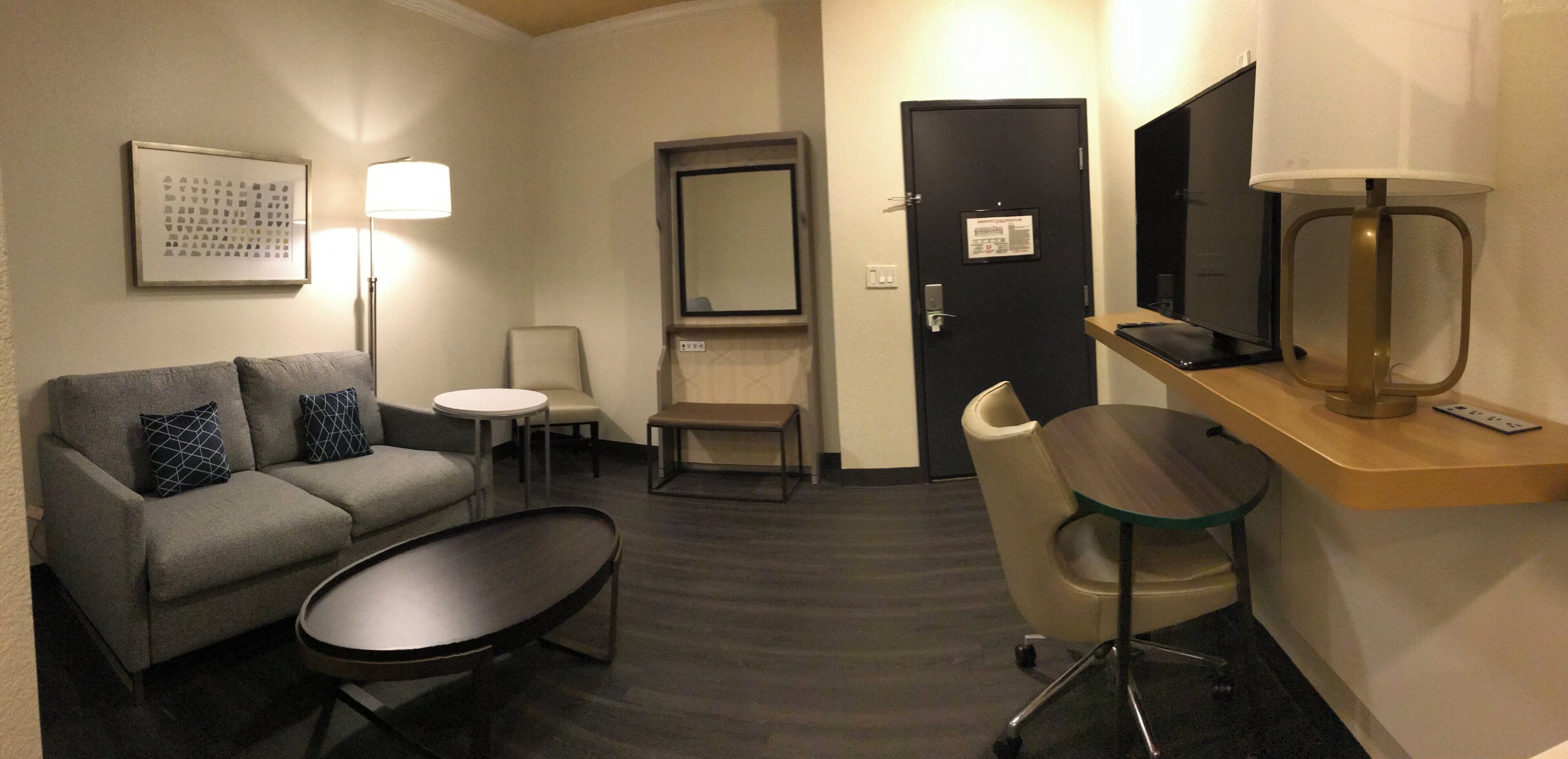 A hotel room with a sofa, a small round table, a desk with a chair, a television, and a lamp.