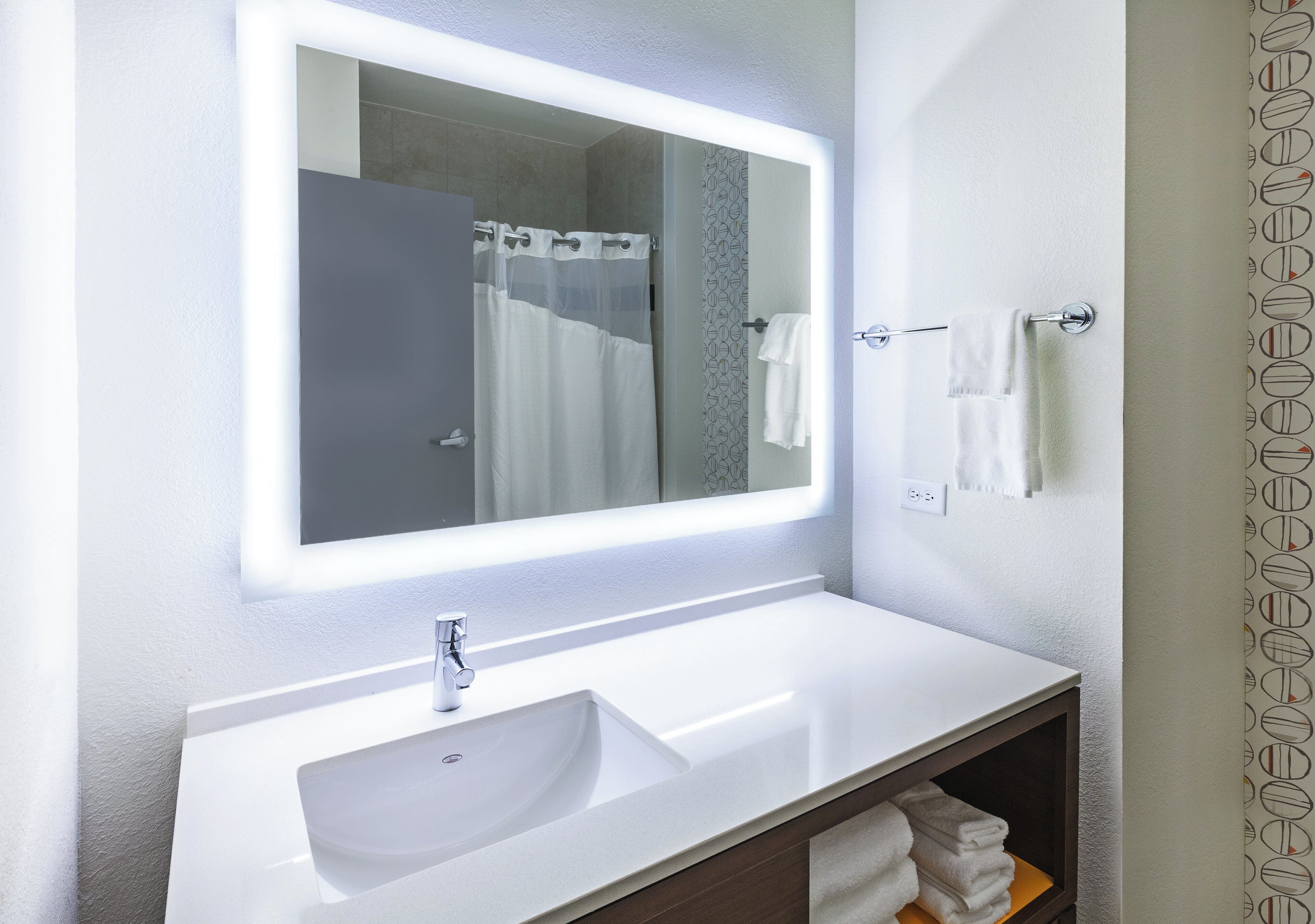 Une salle de bain avec un lavabo blanc, un miroir doté d’un cadre lumineux, un porte-serviettes et un rideau de douche.