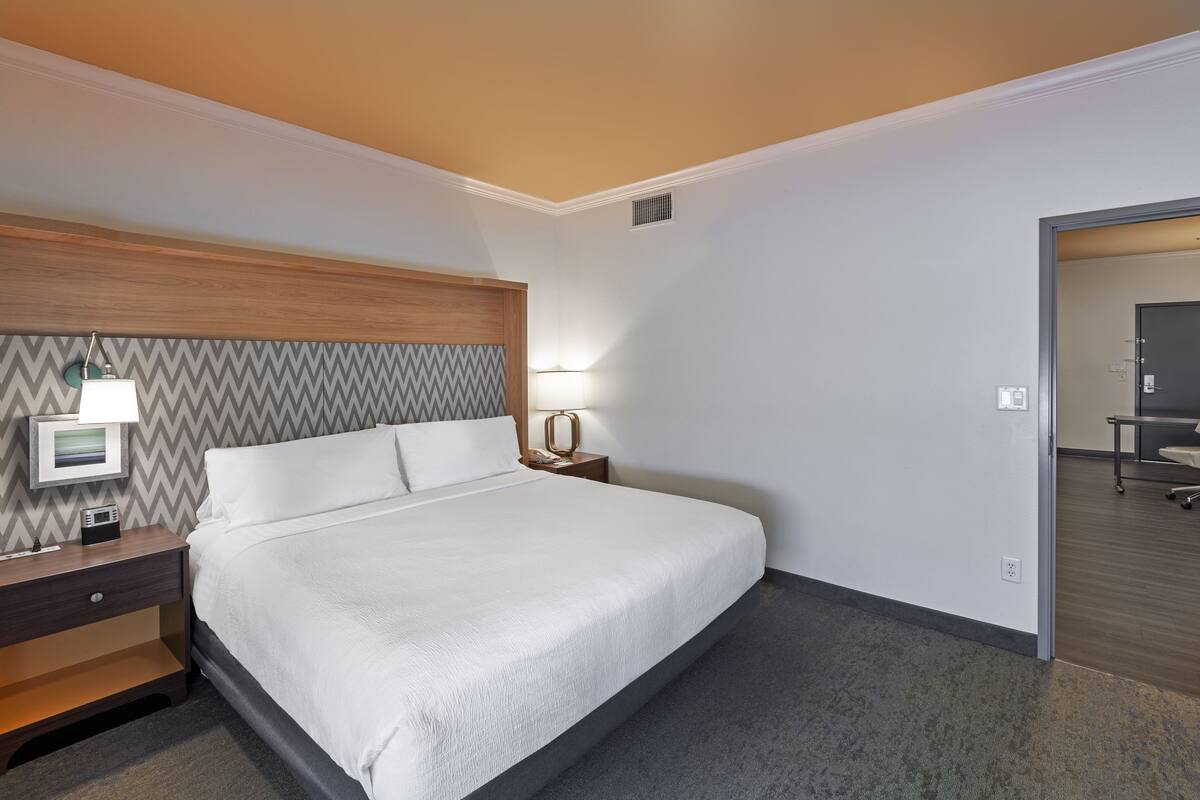 A hotel room with a bed, bedside table, lamp, and a view of the hallway.