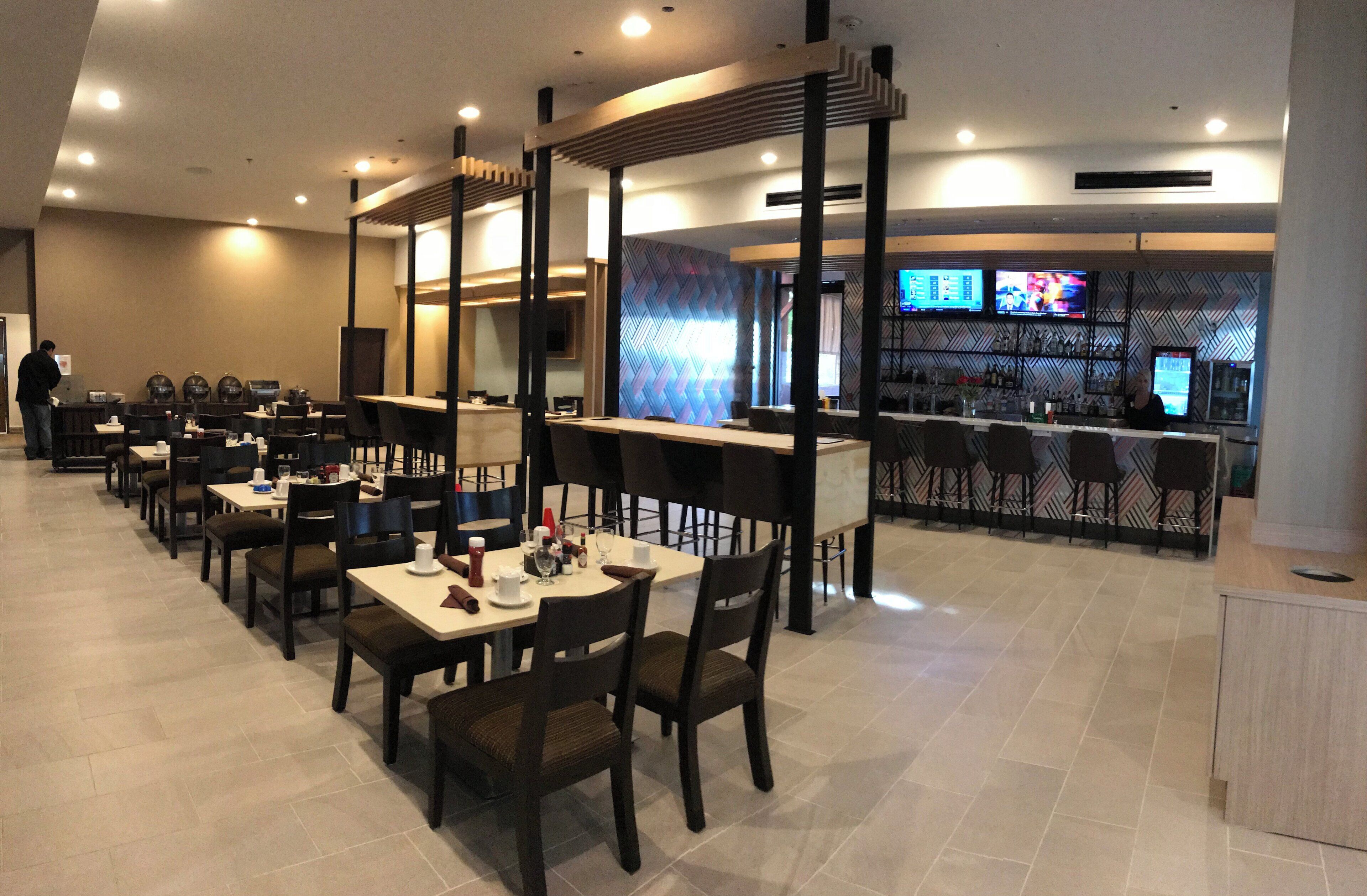 A restaurant with multiple tables and chairs, a bar area with a counter and televisions, and a patterned wall in the background.