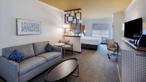 A hotel room with a sofa, a small round table, a bed, a desk with a computer, and a TV.