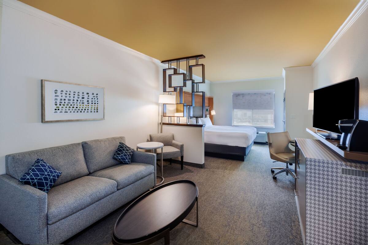 A hotel room with a sofa, a small round table, a bed, a desk with a computer, and a TV.