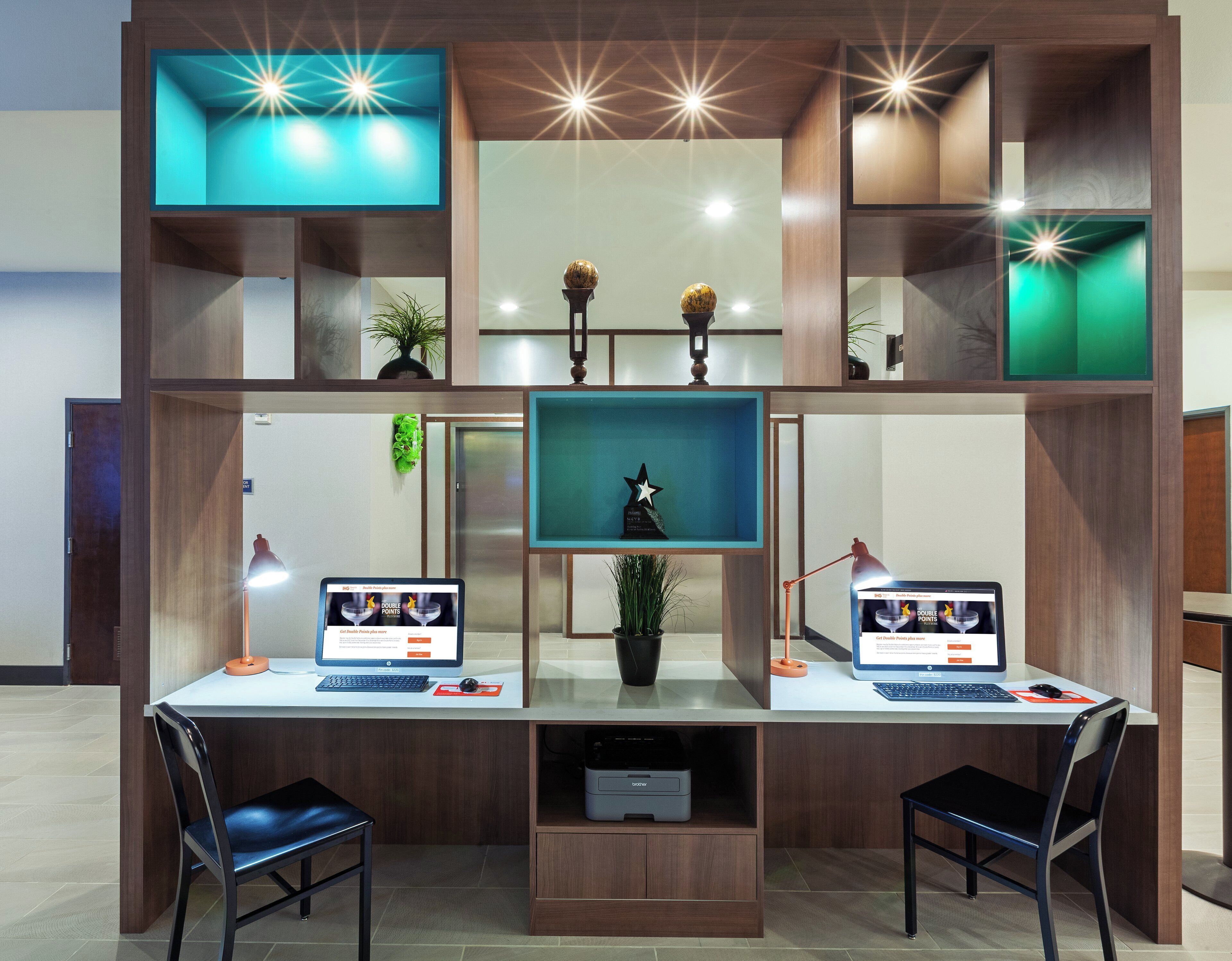 A modern office space with two desks, each with a computer, a lamp, and a chair. There are decorative plants and a trophy on the shelves.