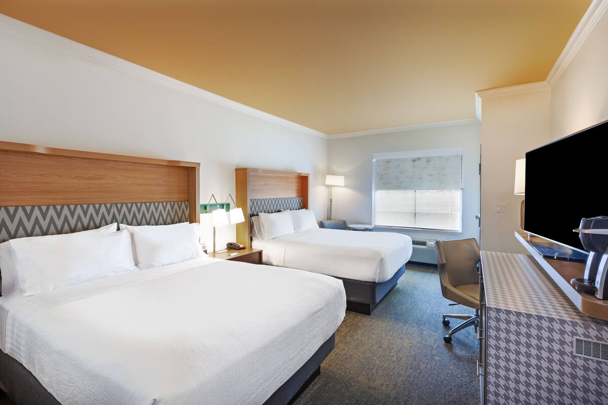 A hotel room with two beds, a desk, a chair, a TV, and a window.