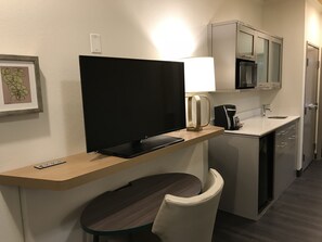 Une chambre d’hôtel équipée d’un téléviseur posé sur un meuble en bois, d’une petite table avec une chaise, d’un coin cuisine avec un four à micro-ondes et d’une machine à café.
