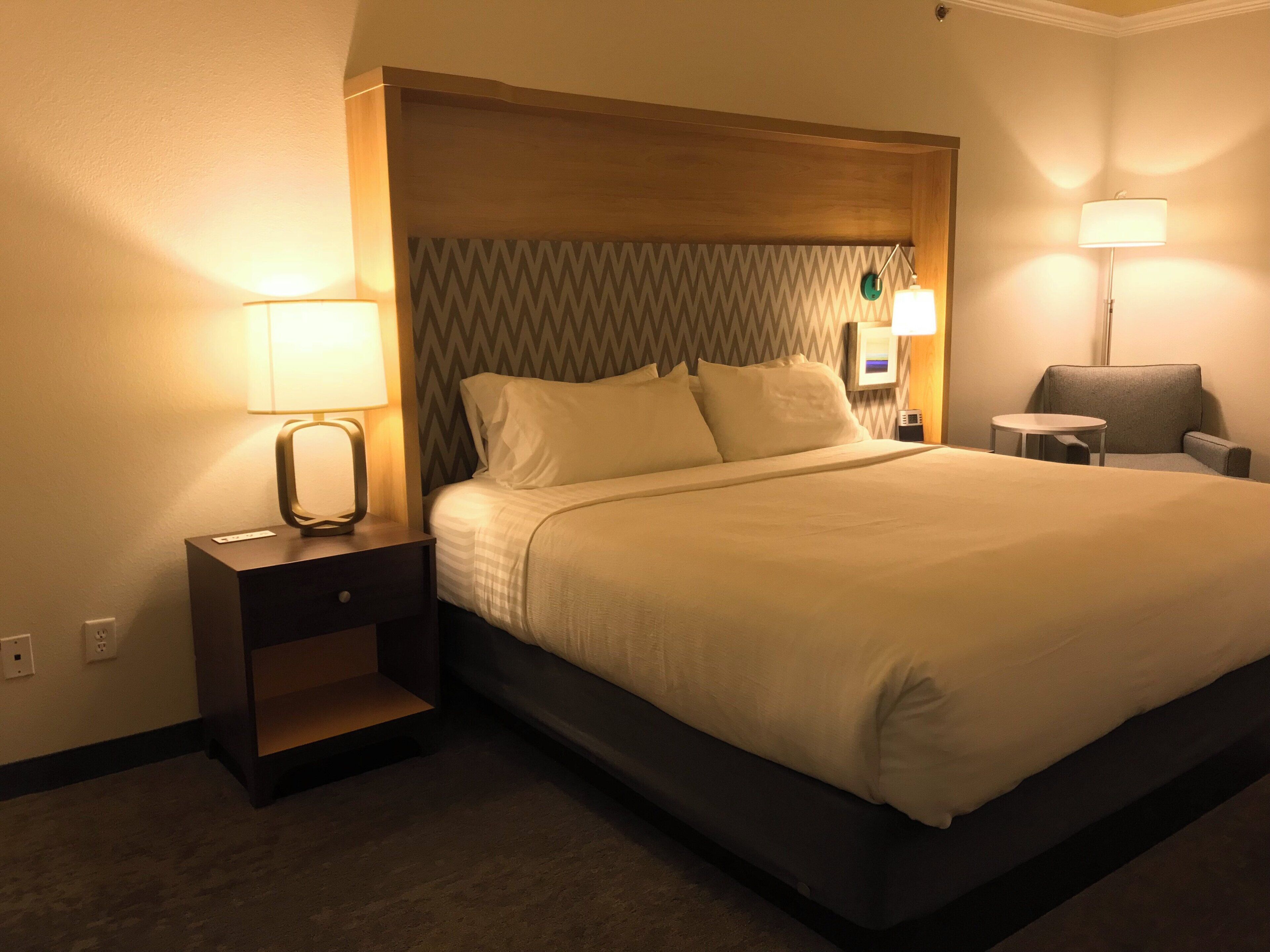 Quarto de hotel com uma cama grande, uma mesa de cabeceira, uma cadeira e duas luminárias.