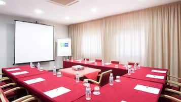 Un salón de conferencias con mesas rojas, botellas de agua y una pantalla para proyector.