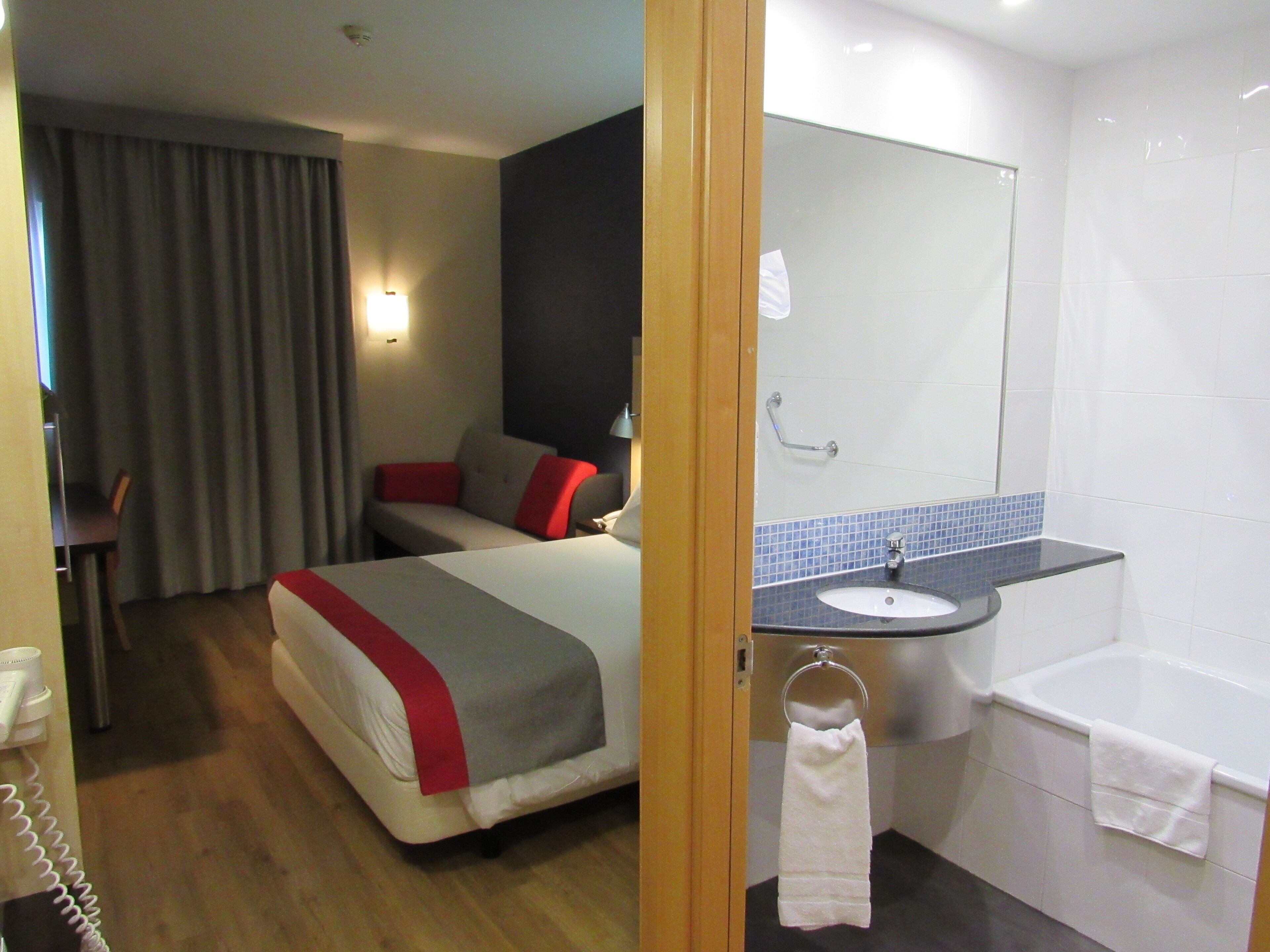 Habitació d'hotel amb un llit, escriptori, cadira, bany amb un vàter i mirall.