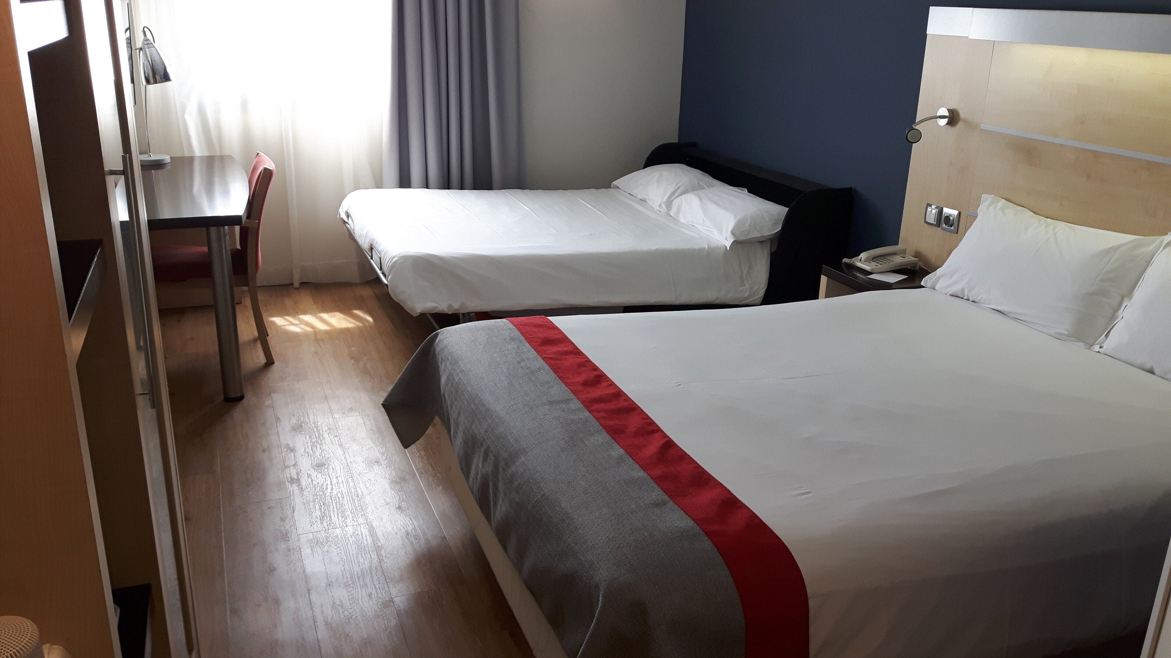 A hotel room with two single beds, a desk, a chair, and a bedside telephone.