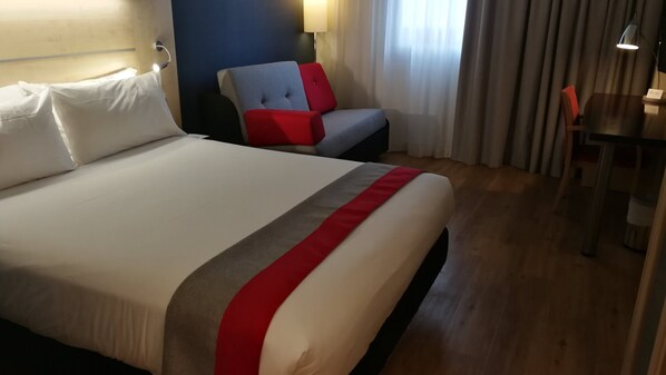 A hotel room with a large bed, a chair with red cushions, a desk, and a lamp.