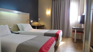 A hotel room with two beds, a desk, a television, and a window with curtains.