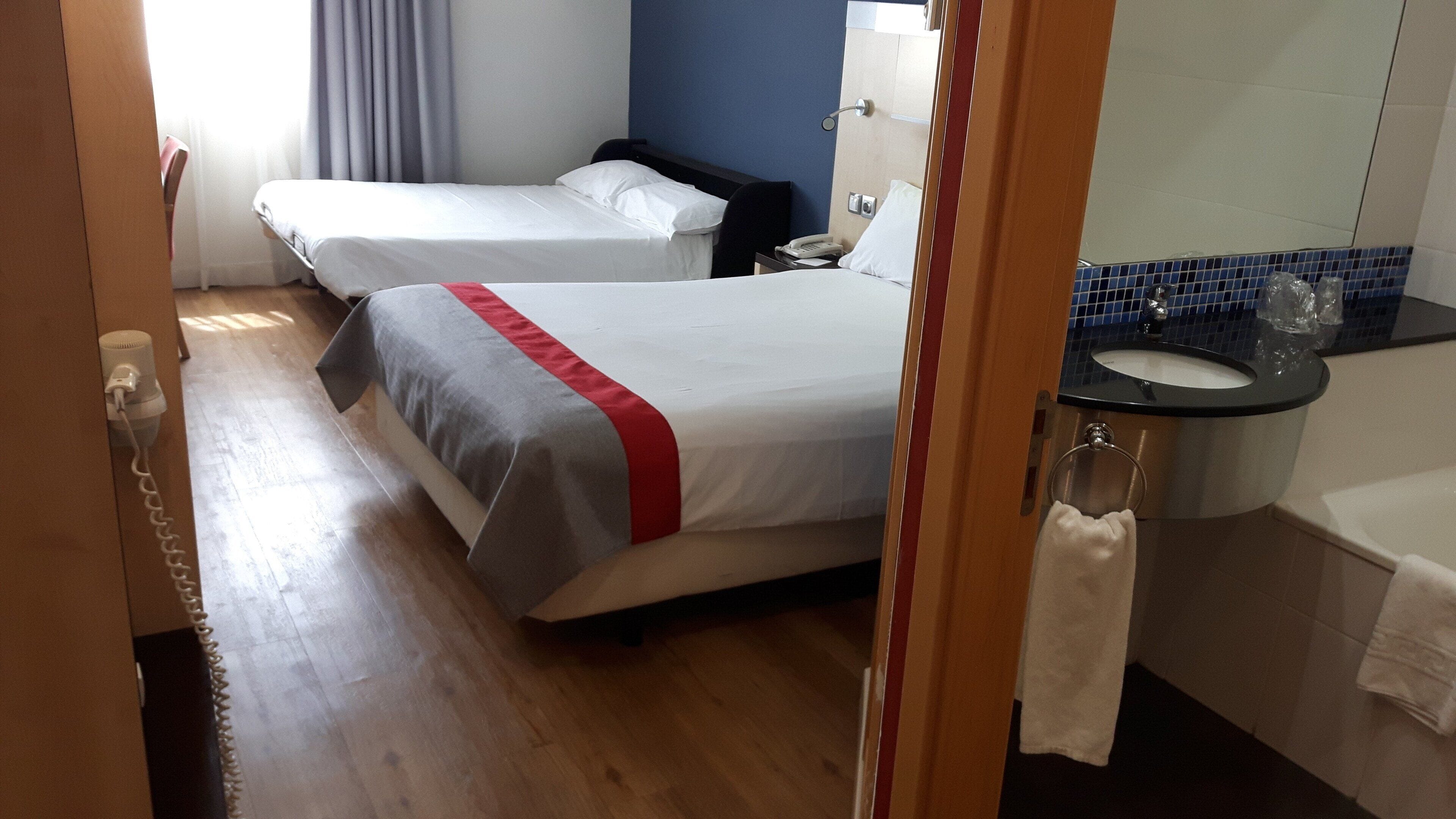 Habitación de hotel con dos camas, baño con lavamanos y espejo, y piso de madera.