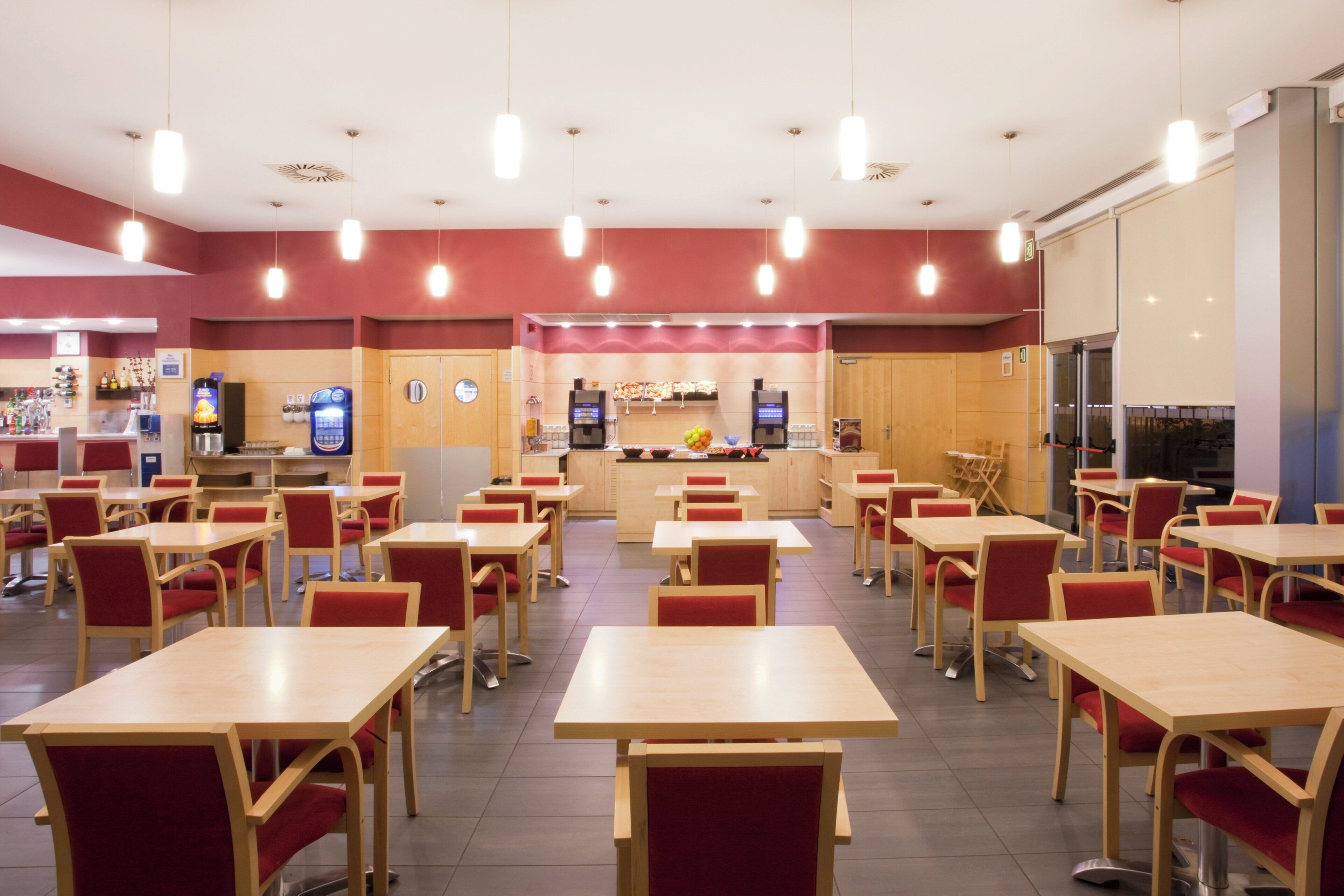 A cafeteria with multiple tables and chairs, a counter with a beverage dispenser, and a kitchen area in the background.