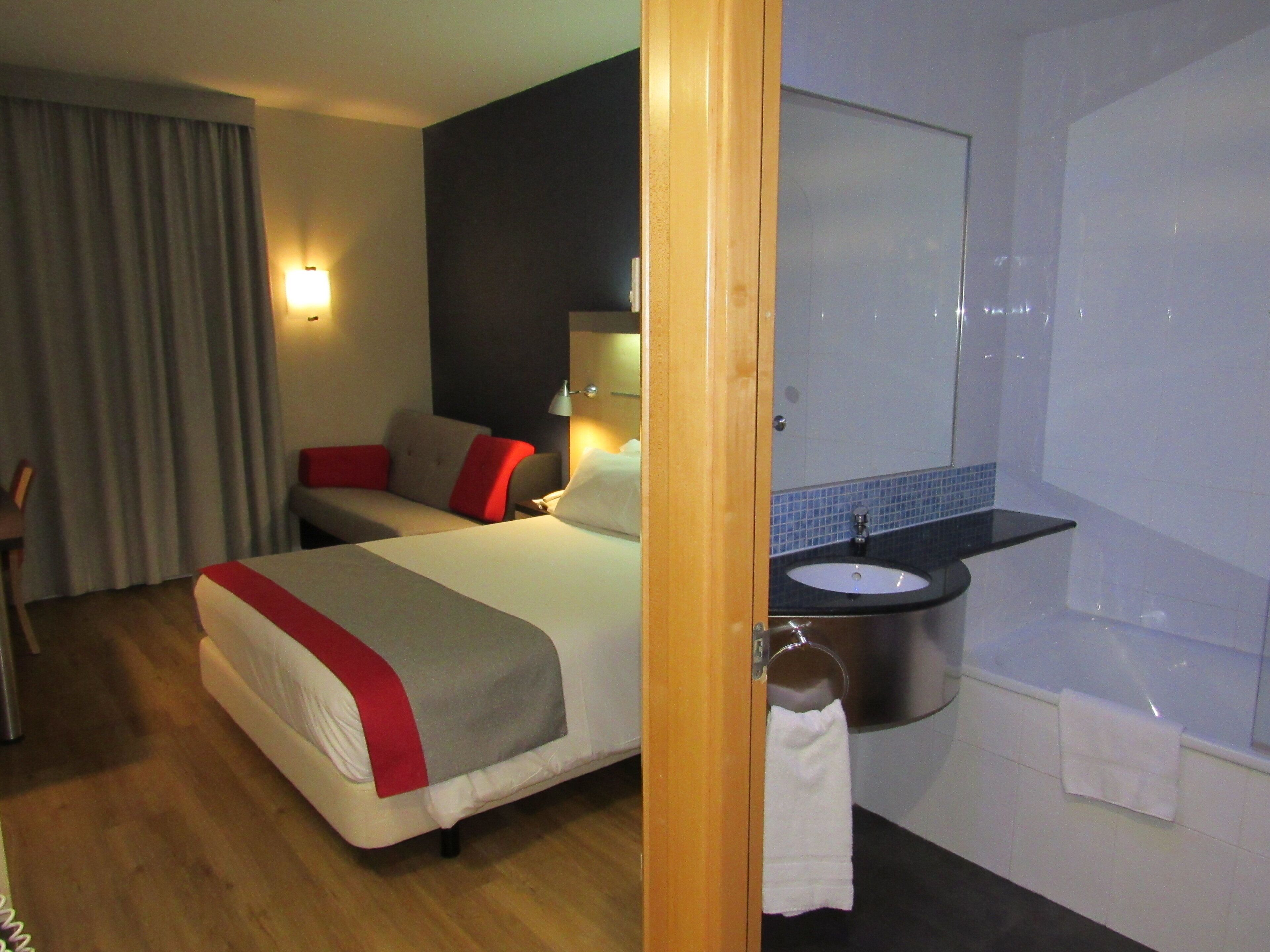 A hotel room with a bed, a bedside table, a lamp, a bathroom with a sink and mirror, and a towel rack.