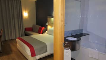 Habitación de hotel con cama, mesita de noche, lámpara, baño con lavamanos y espejo, y un perchero para toallas.