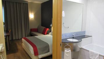 A hotel room with a bed, a desk, a chair, a bathroom with a sink, a mirror, and a bathtub.