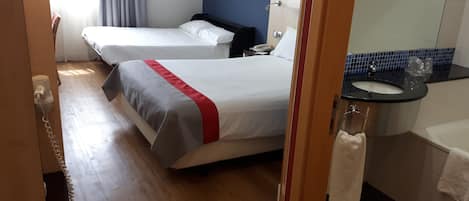 A hotel room with two beds, a bathroom with a sink and mirror, and a wooden floor.