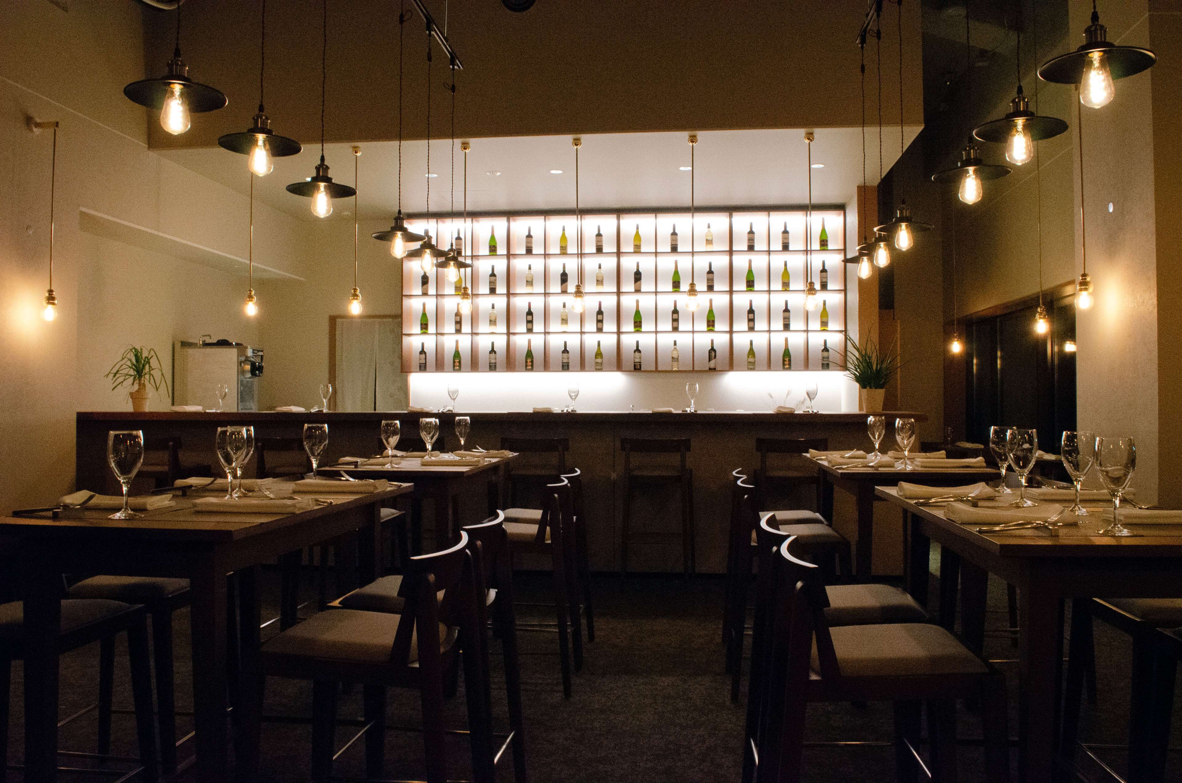 Um restaurante com várias mesas, cadeiras, luminárias pendentes e uma parede de garrafas de vinho.