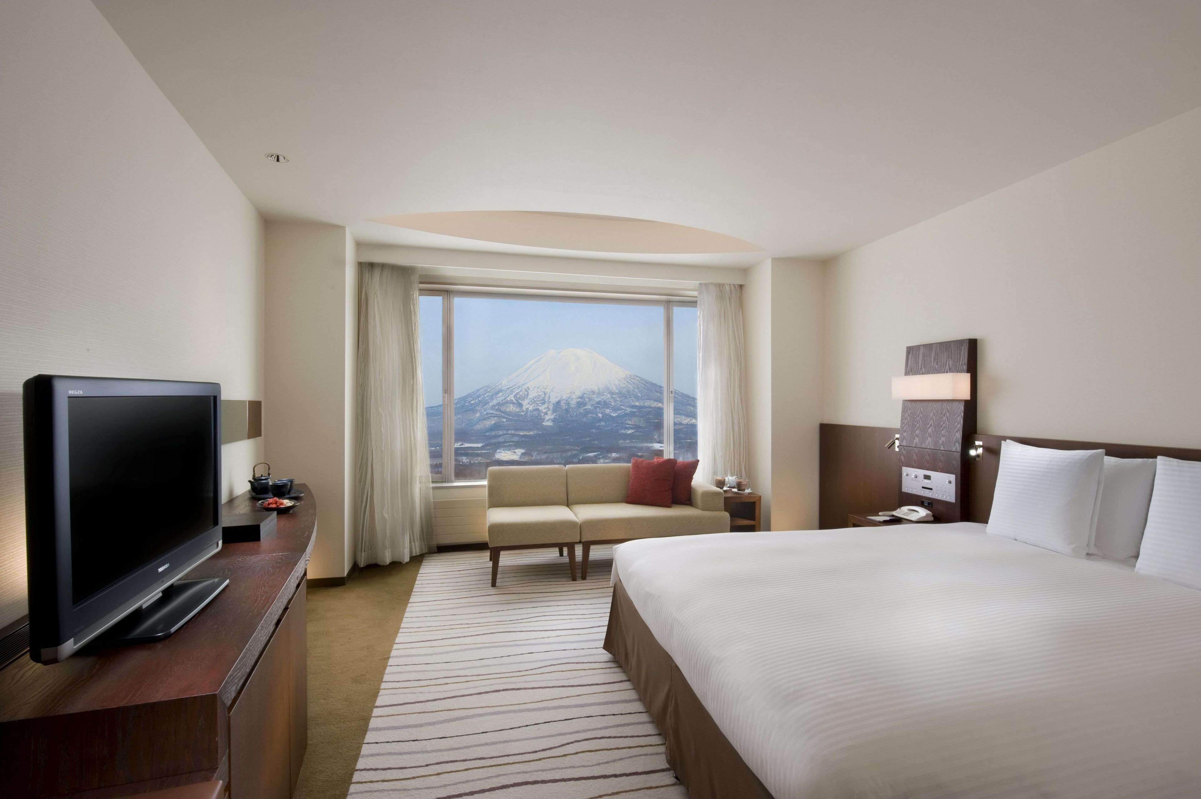 Habitación de hotel con cama, televisión, escritorio, silla, sofá, vista a la montaña y mesita de noche con teléfono.