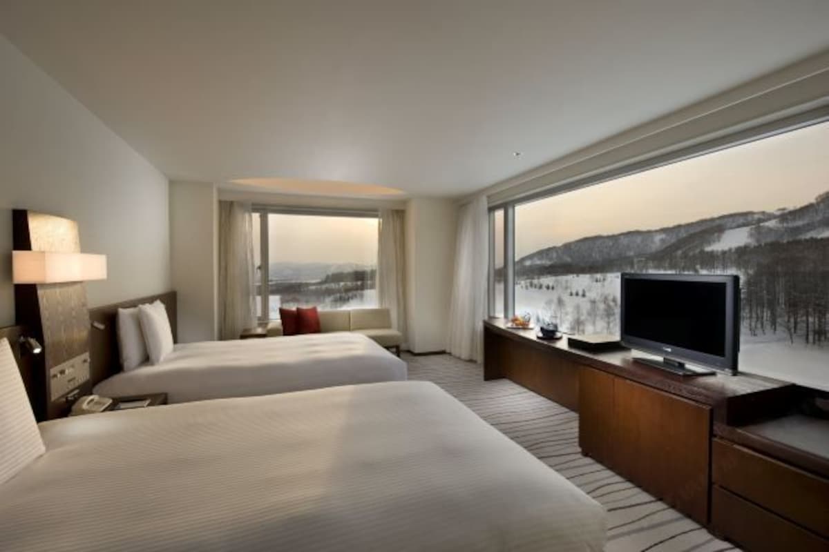 Une chambre d’hôtel avec deux lits, une télévision, un bureau et une grande fenêtre donnant sur un paysage enneigé.