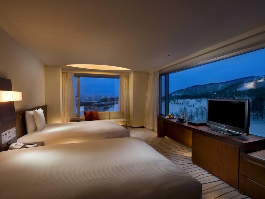 A hotel room with two beds, a TV, a desk, and a large window with a mountain view.