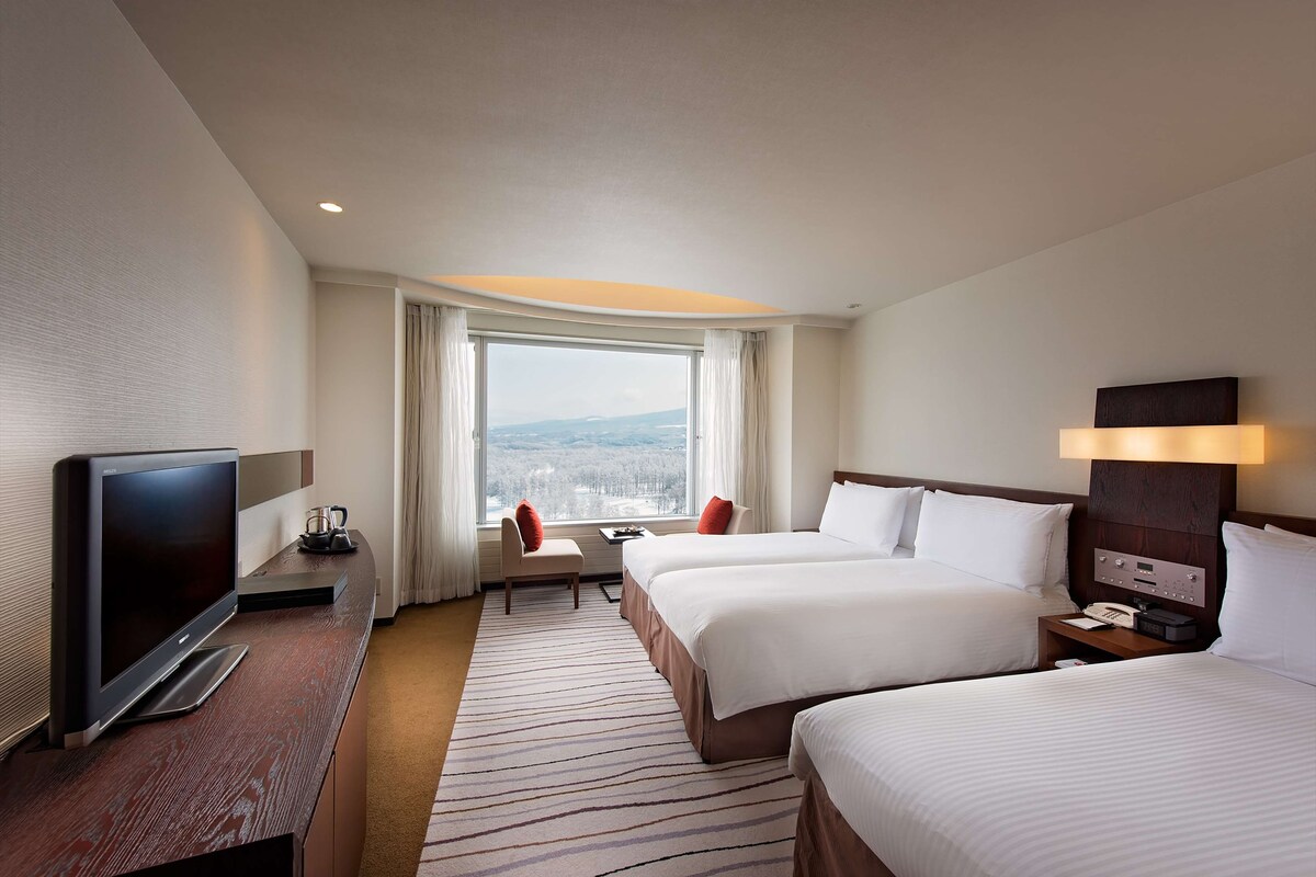 A hotel room with two beds, a TV, a desk, and a window with a view.