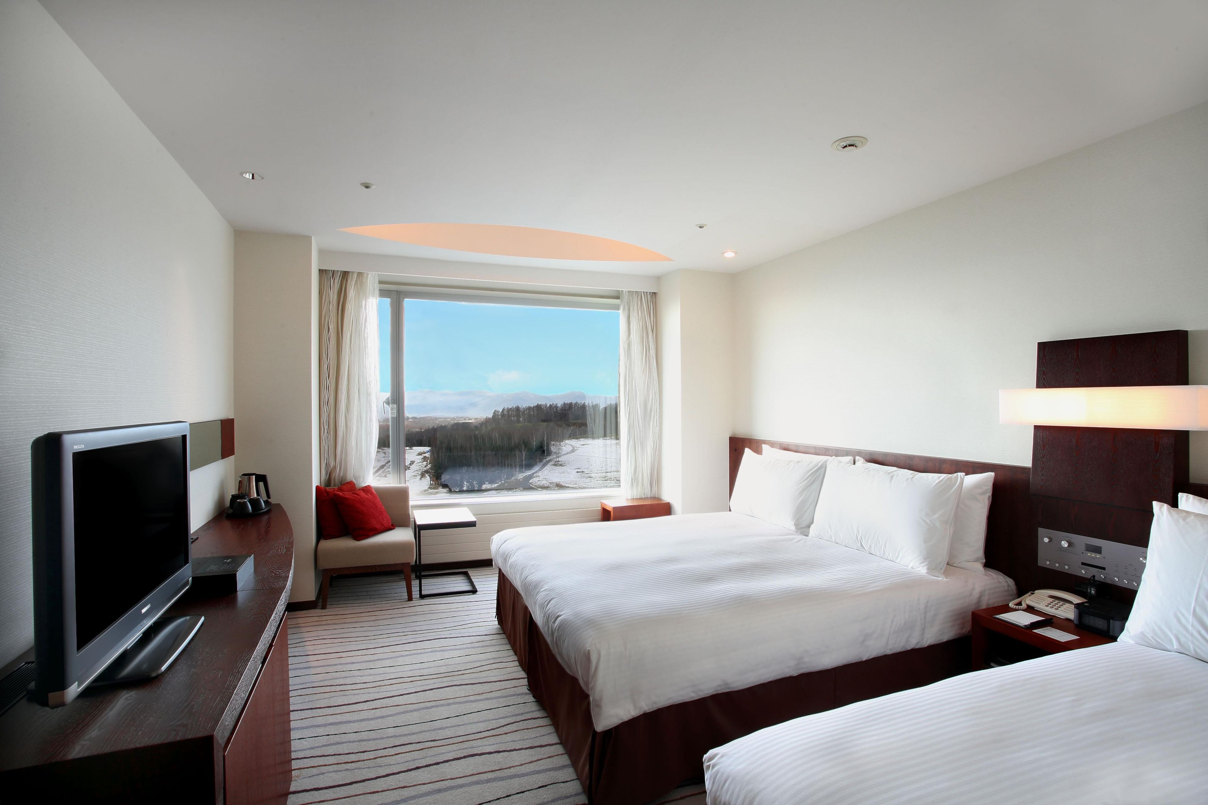 Habitación de hotel con dos camas, televisor, escritorio y ventana con vista a un paisaje nevado.