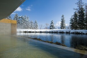 Uma área de spa com uma nascente termal, rodeada por árvores cobertas de neve e uma montanha ao fundo.