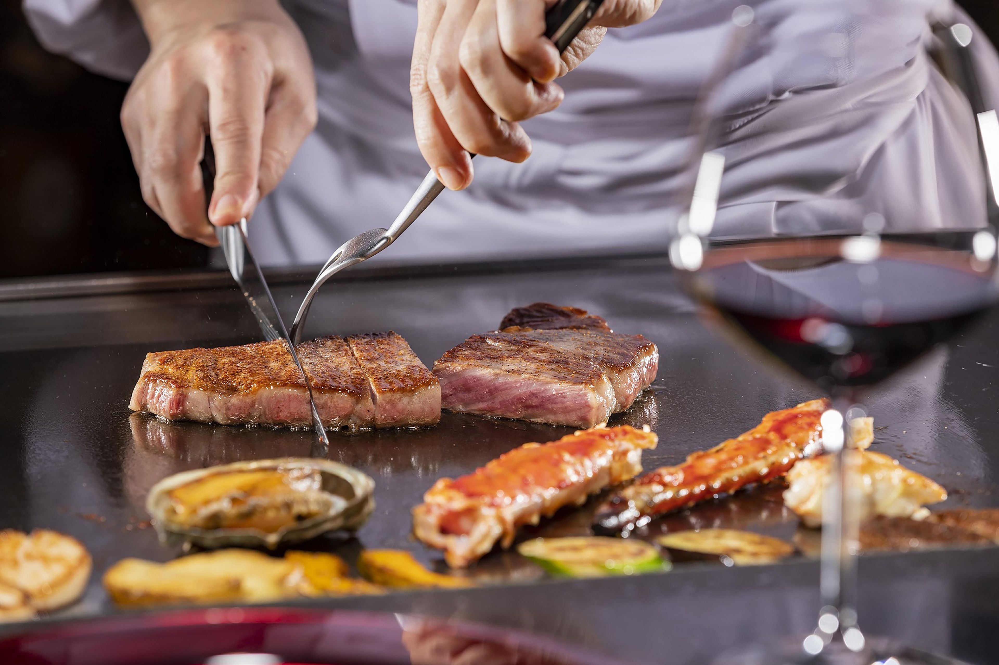 A person slicing a steak on a grill with vegetables and a glass of red wine nearby.