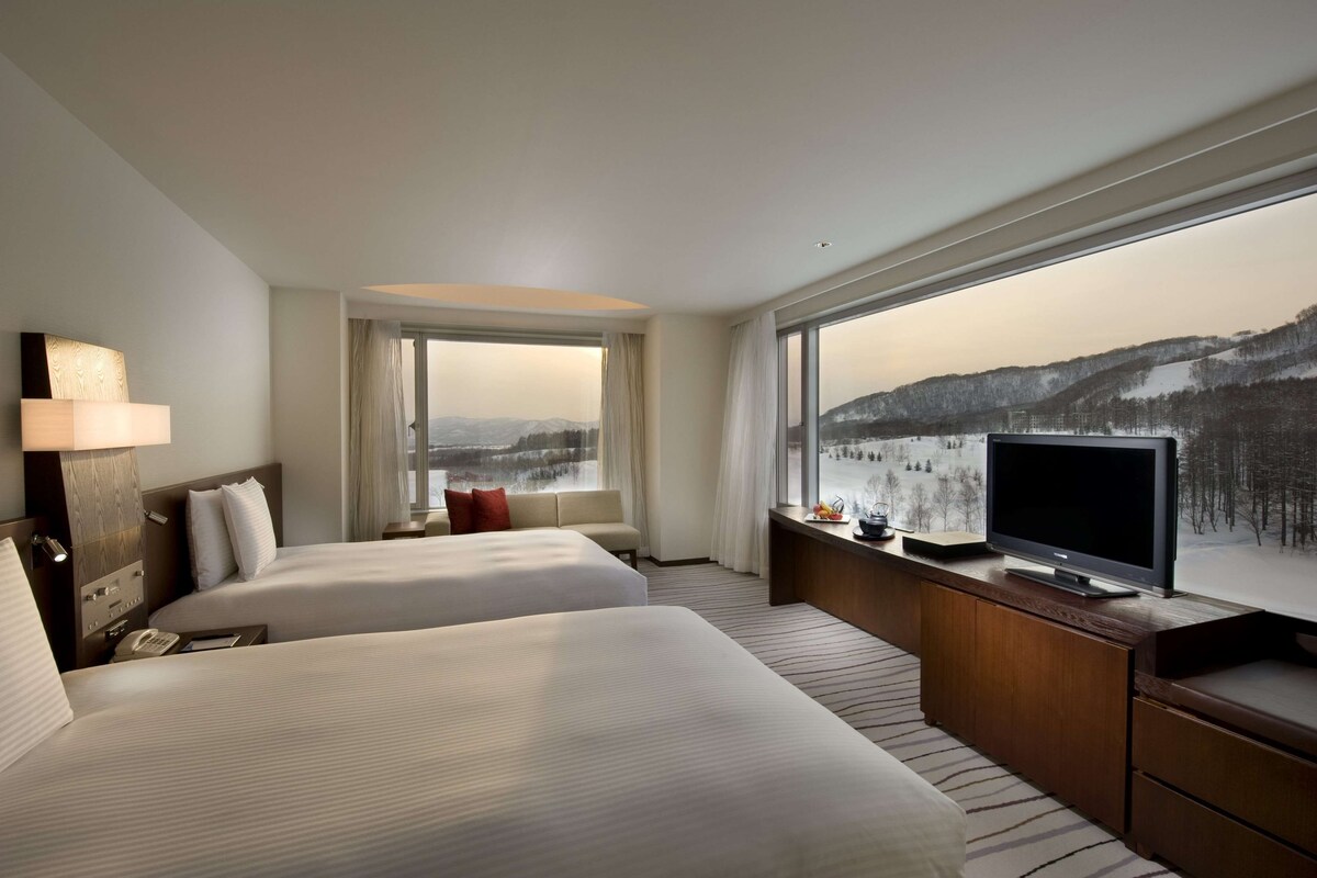 A hotel room with two beds, a TV, a large window with a snowy landscape view, and a wooden dresser.