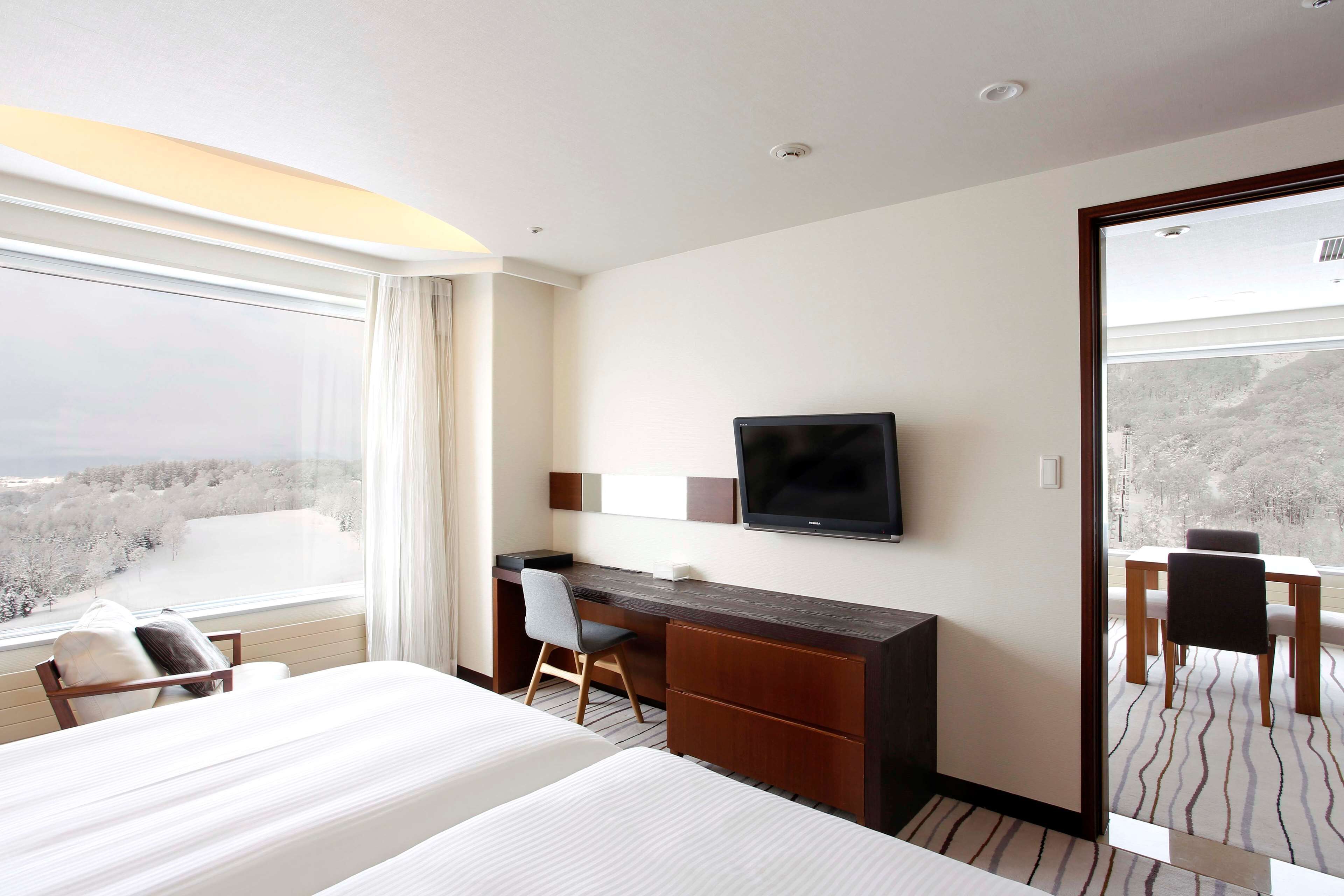 A hotel room with a bed, a desk, a chair, a television, and a large window offering a view of the city.