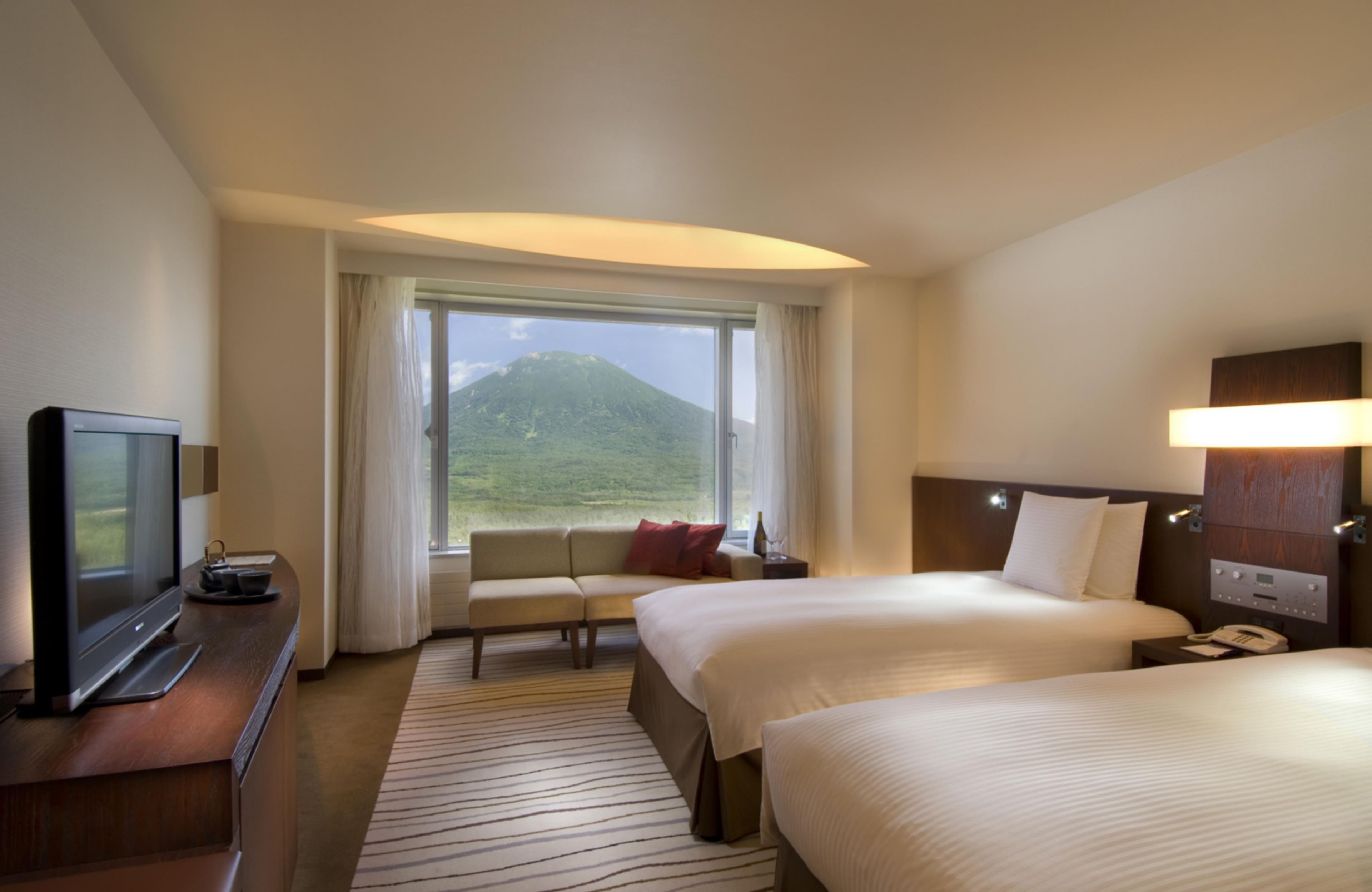 A hotel room with a bed, a TV, a desk, and a window with a mountain view.