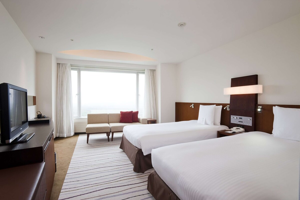 A hotel room with two beds, a TV, a sofa, and a large window with curtains.