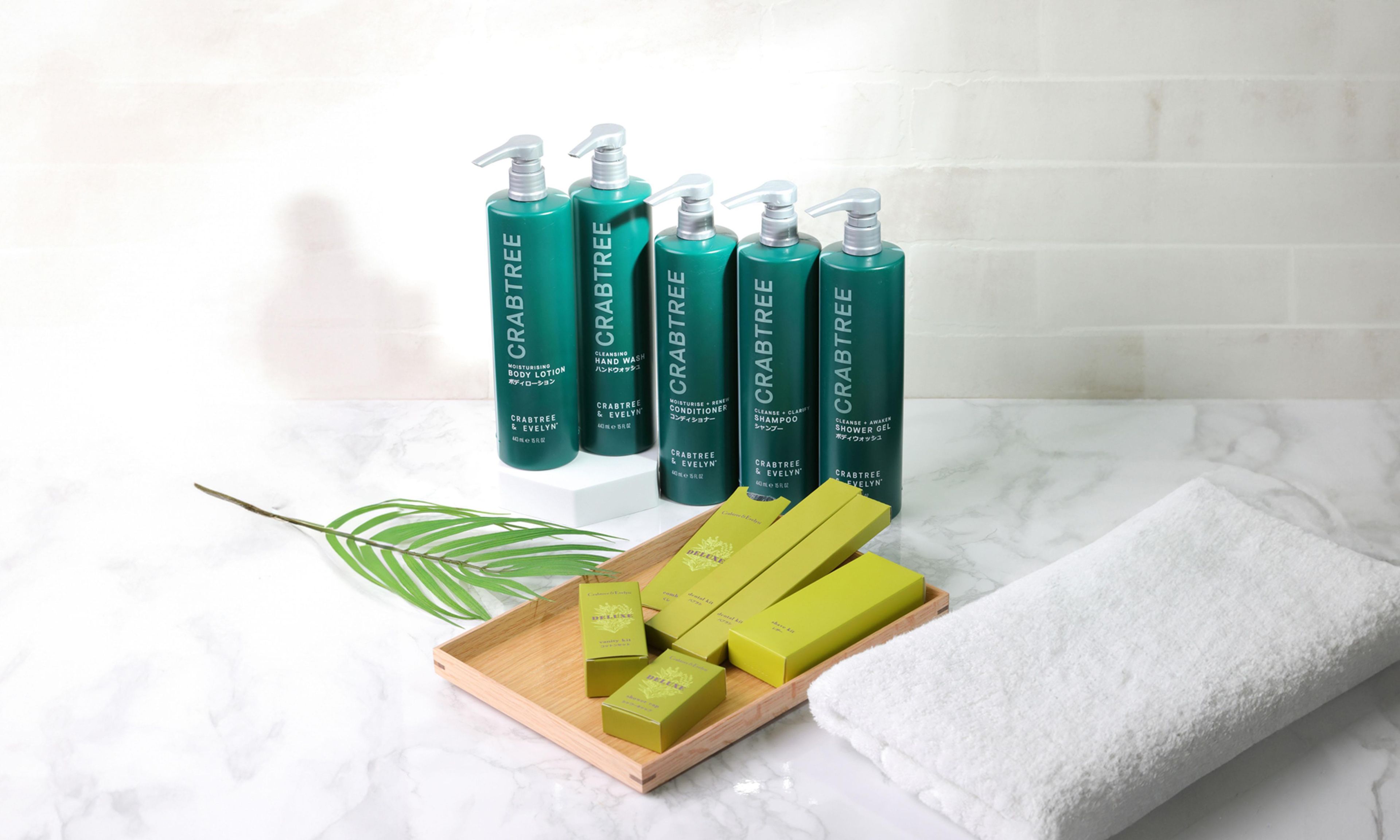 A set of green bottles with pump dispensers, arranged on a marble surface with a white towel and a wooden tray.