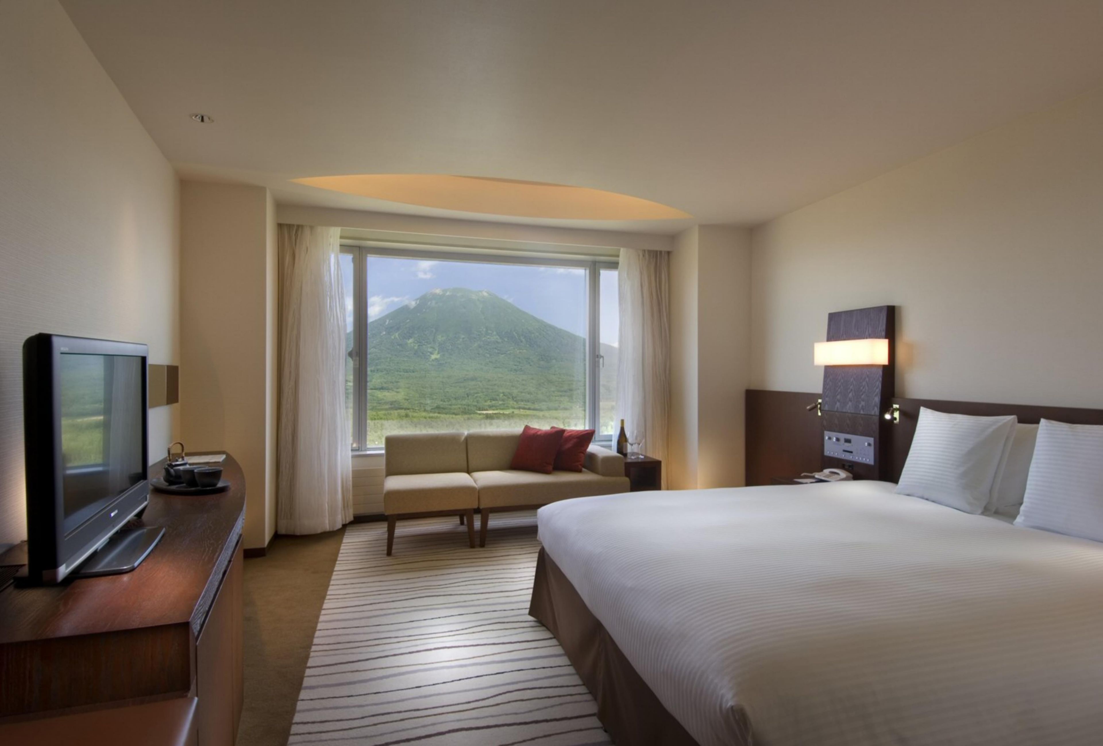 A hotel room with a large bed, a TV, a desk, a window with a mountain view, and a nightstand.