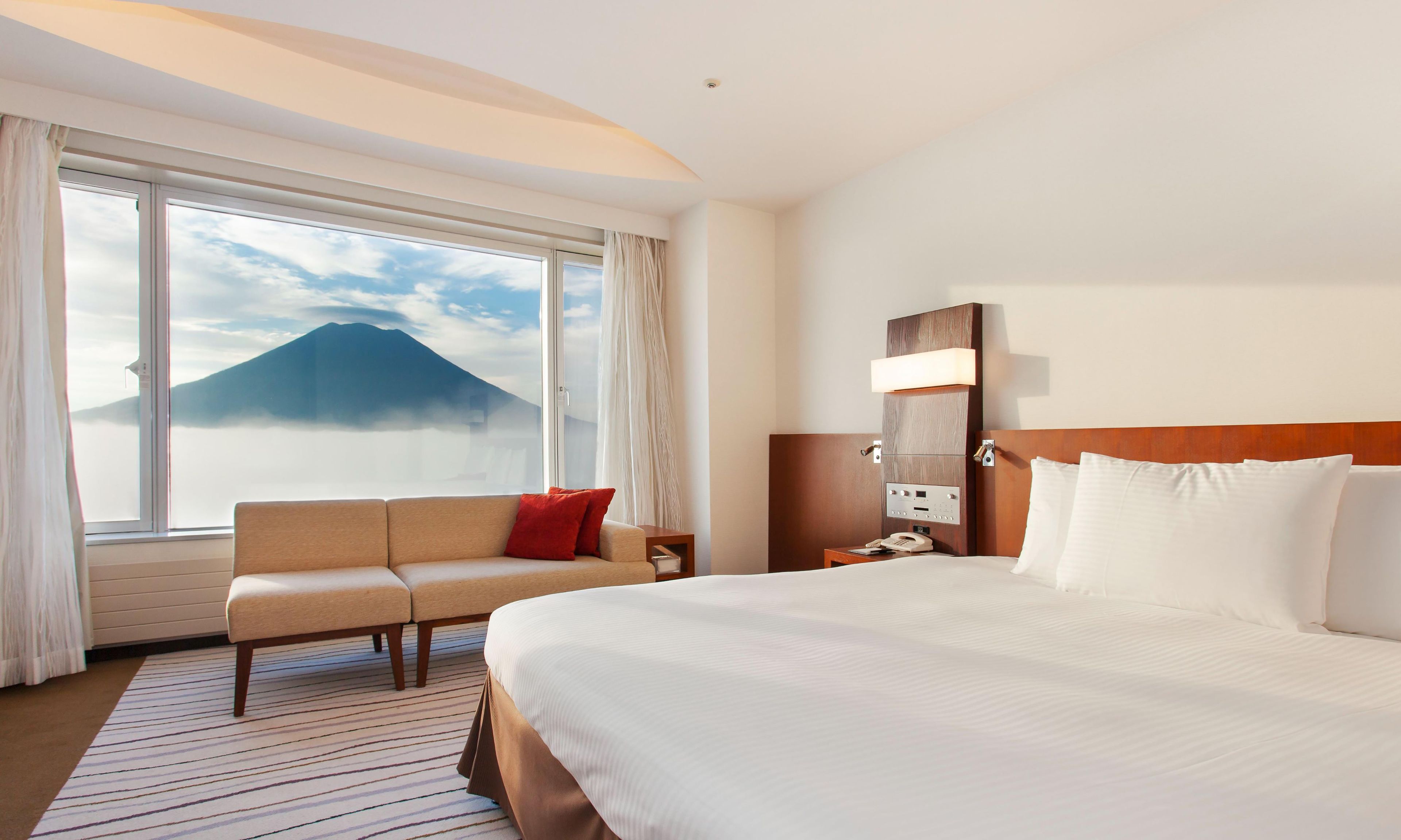 Habitación de hotel con cama, sofá, mesita de noche, lámpara y ventana con vista a la montaña.