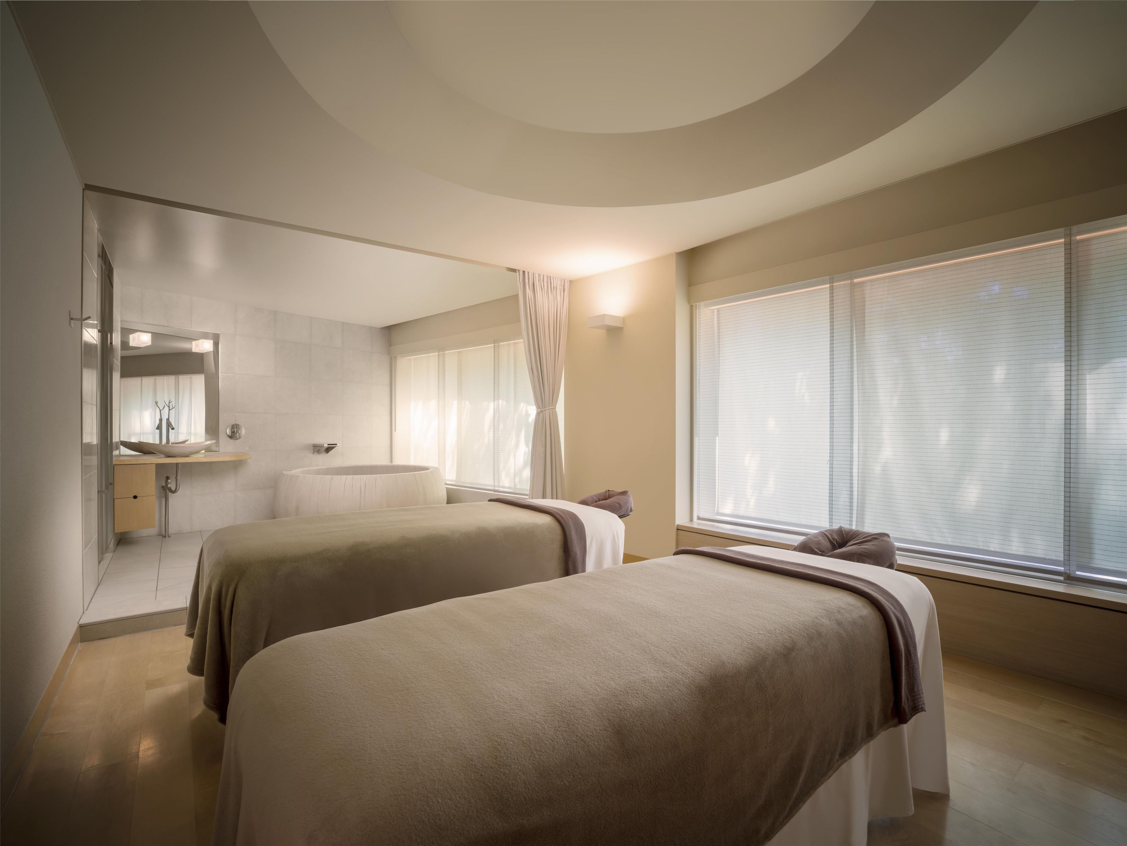 A modern hotel room with two beds, a large window with blinds, a bathroom with a bathtub, and a mirror above the sink.