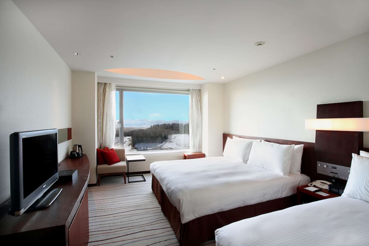 Ein Hotelzimmer mit zwei Betten, einem Fernseher, einem Schreibtisch und einem Fenster mit Blick auf eine verschneite Landschaft.