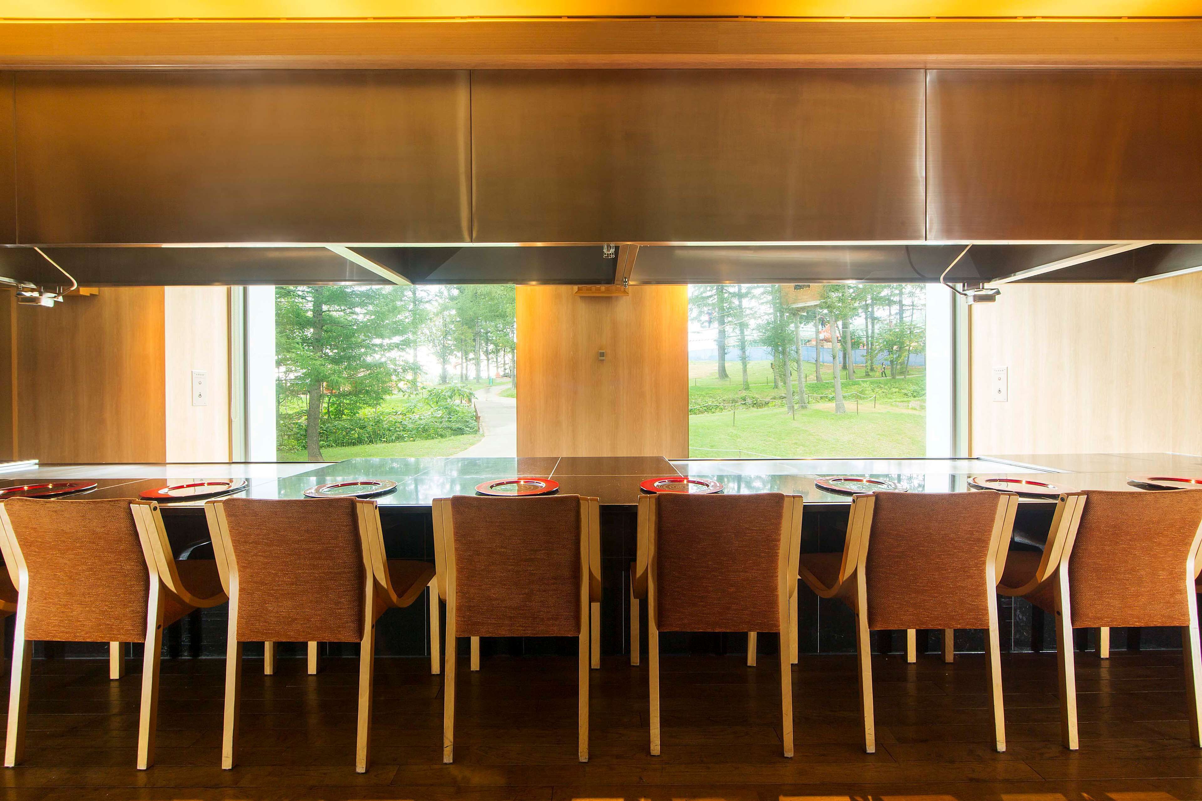 A modern dining area with a long table, six chairs, and a view of a green landscape through large windows.